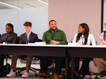 UM student talking at a conference table while other students look on.